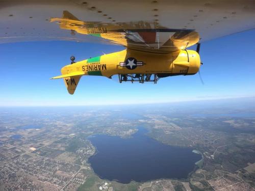 A yellow biplane flying upside down over a large lake and landscape under a clear blue sky.