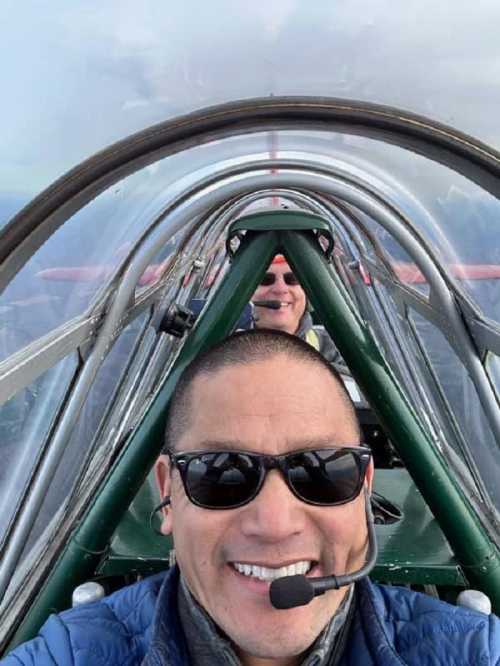 Two pilots take a selfie inside a small aircraft, wearing sunglasses and headsets, with a clear sky in the background.