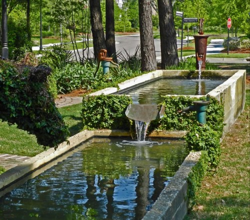 A serene landscape featuring a water feature with flowing water, surrounded by greenery and trees along a quiet street.
