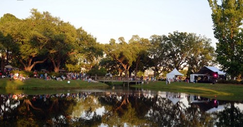 A lively outdoor event by a pond, with people gathered near a stage and trees reflecting in the water.