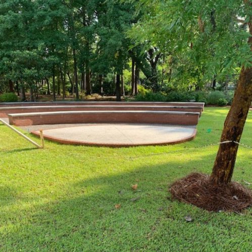 A circular concrete platform surrounded by grass and trees, with wooden seating along the edge.