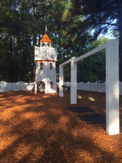 A playground featuring a white tower with an orange roof and swings, surrounded by wood chips and a white fence.