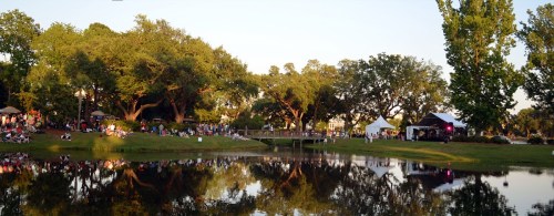 A serene park scene with a pond reflecting trees and a crowd enjoying an outdoor event near a stage.