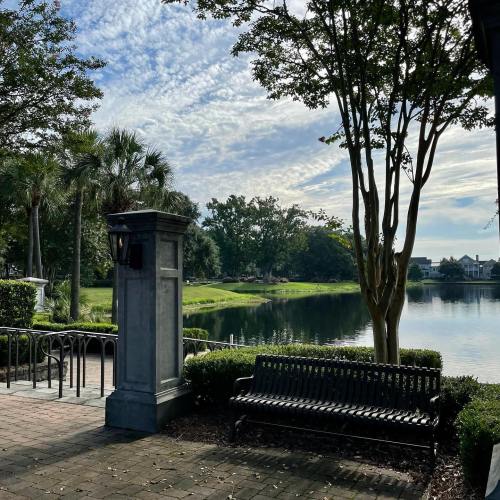 A serene park scene featuring a bench by a calm lake, surrounded by trees and a blue sky with scattered clouds.