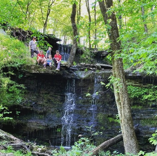 Three people sit on a rocky ledge by a small waterfall, surrounded by lush green trees and foliage.