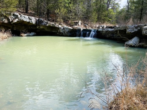 A serene pond surrounded by rocky terrain and trees, featuring a small waterfall cascading into the water.