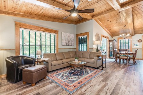 Cozy living room with a leather sofa, wooden ceiling, and dining area, featuring a rug and large windows with blinds.