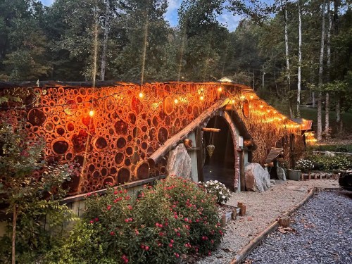 A unique, rustic building with a wooden facade, illuminated by warm lights, surrounded by trees and flowers.