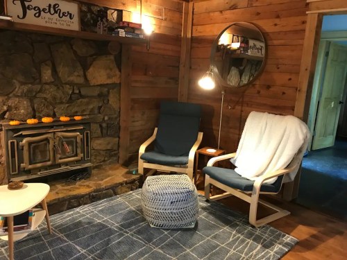 Cozy living room with a stone fireplace, two chairs, a round mirror, and a patterned rug. Warm, inviting atmosphere.