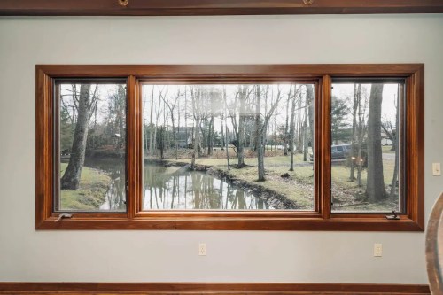 A large window with a wooden frame overlooks a serene outdoor scene with trees and a calm waterway.