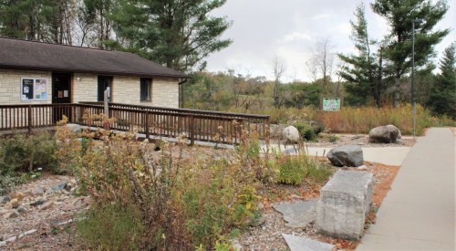 A stone building with a wooden deck surrounded by greenery and rocks, set against a cloudy sky.