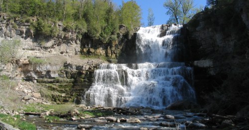 A cascading waterfall flows down rocky cliffs, surrounded by lush greenery and a clear blue sky.