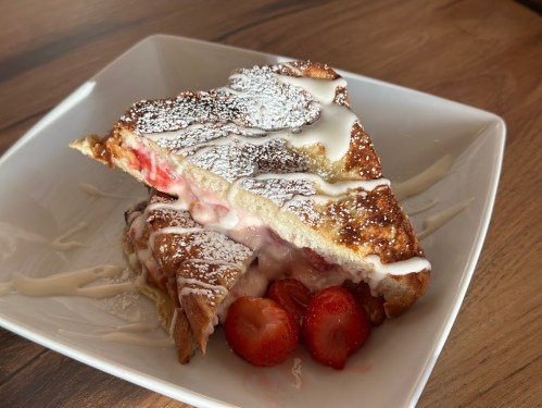 A slice of stuffed French toast with strawberries, drizzled with icing and dusted with powdered sugar on a white plate.