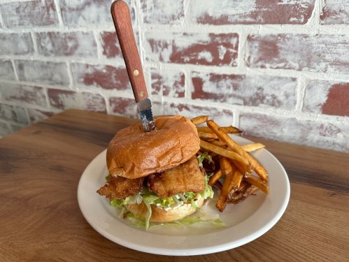 A crispy fish sandwich with lettuce on a plate, accompanied by golden fries and a knife stuck in the bun.