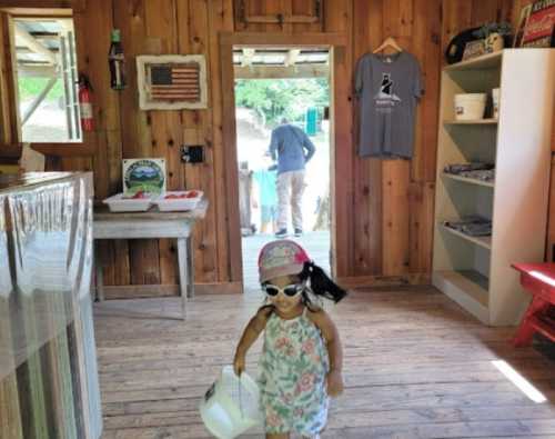 A child in sunglasses and a hat walks inside a wooden cabin, carrying a basket, with adults visible in the background.