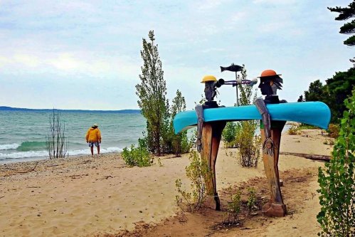 Two wooden figures with canoes stand on a sandy beach, while a person walks along the shore by the water.