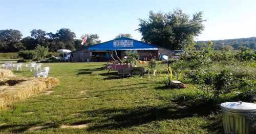 A rustic farm stand with a blue roof, surrounded by green grass, hay bales, and chairs under a sunny sky.