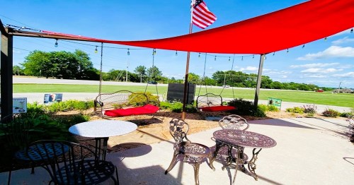 Outdoor seating area with red shade sails, swings, and tables, set against a sunny sky and green landscape.