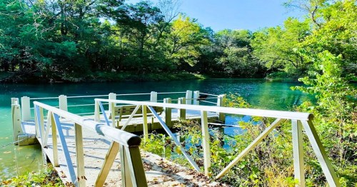 A wooden dock extends over a serene, blue-green lake surrounded by lush greenery and trees under a clear sky.