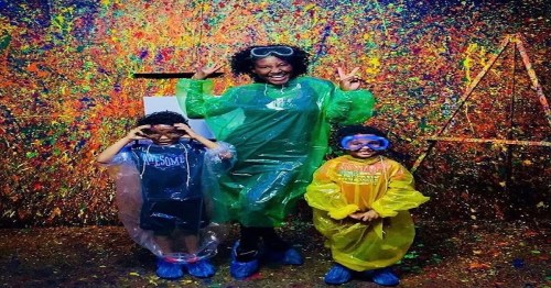Three children in colorful ponchos pose playfully in front of a vibrant, splattered paint backdrop.
