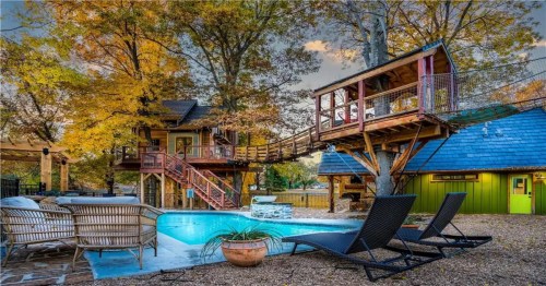 A treehouse with a deck overlooks a pool surrounded by autumn trees and outdoor seating.