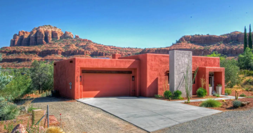 A modern adobe-style house with a garage, set against a backdrop of red rock formations and greenery.