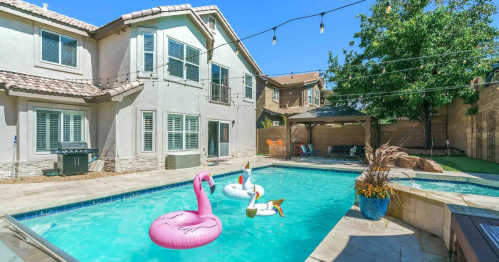 A backyard pool with pink flamingo and swan floats, surrounded by a house, trees, and string lights.