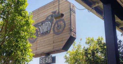 Sign featuring a vintage motorcycle design, hanging above a building, surrounded by green trees and a clear blue sky.