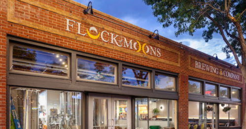 Exterior of Flock & Moons Brewing Company, featuring a brick facade and large windows, under a blue sky.