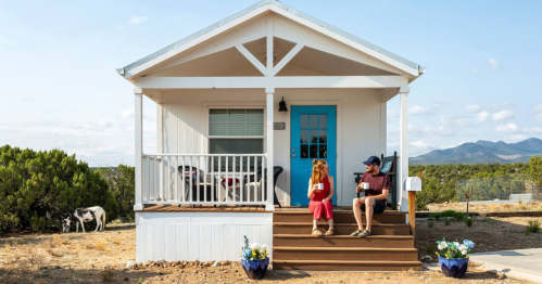 A cozy white house with a blue door, featuring a person sitting on the steps and a cow in the background.