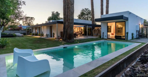 Modern backyard with a swimming pool, palm trees, and two white chairs, surrounded by a well-maintained garden.