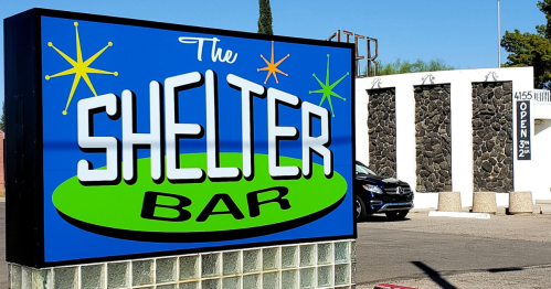 Bright sign for "The Shelter Bar" with a retro design, featuring stars and a green background, next to a stone wall.