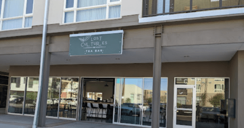 Exterior of "Lost Cultures Tea Bar" with large windows and a sign above the entrance. Modern building design.