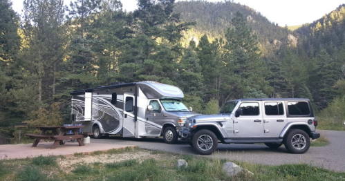 A camper van parked beside a silver Jeep in a forested area with mountains in the background.
