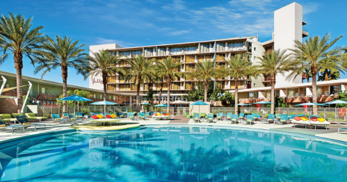 A sunny pool area with palm trees, lounge chairs, and a colorful hotel in the background. Clear blue sky above.