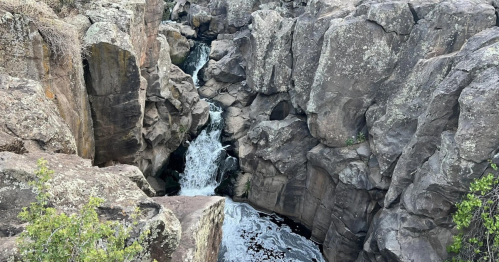 A narrow waterfall cascades through rocky cliffs, surrounded by rugged terrain and sparse greenery.