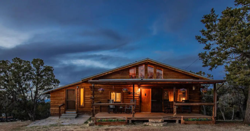 A cozy wooden cabin at dusk, with warm lights glowing from the windows and surrounded by trees.