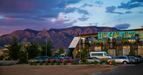 A colorful sunset over mountains, with a modern building and parked cars in the foreground.