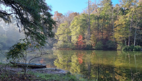 A serene lake surrounded by trees with autumn foliage, featuring a small boat on the shore and morning mist.