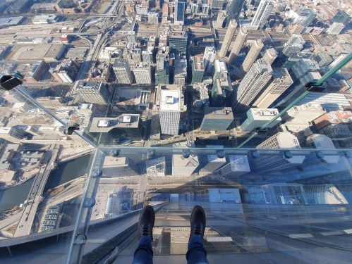 A person stands on a glass floor overlooking a cityscape with tall buildings and streets below.