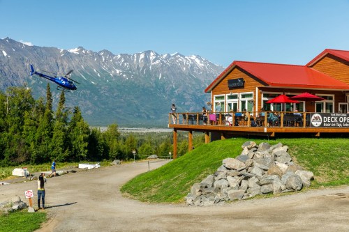 A helicopter flies near a mountain lodge with outdoor seating, surrounded by lush greenery and rocky terrain.