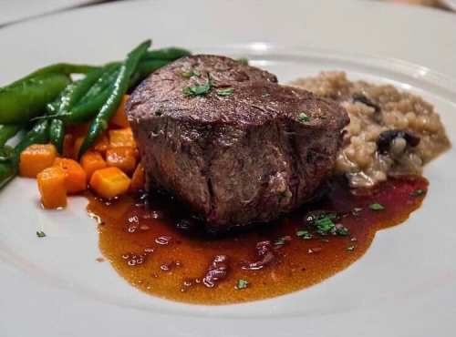 A plated meal featuring a beef filet, green beans, carrots, and mushroom risotto drizzled with sauce.