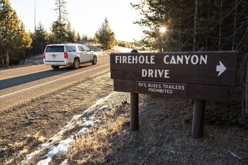 A sign for Firehole Canyon Drive beside a road, with a vehicle passing and trees in the background.