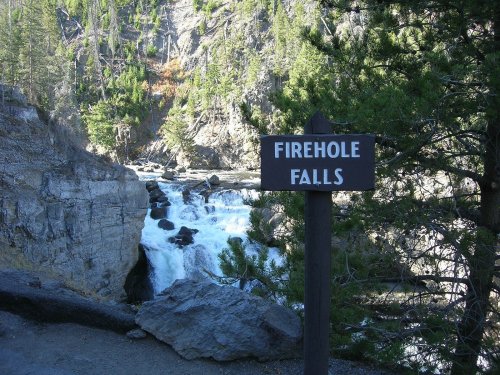 Sign for Firehole Falls beside a rushing waterfall, surrounded by rocky cliffs and lush greenery.