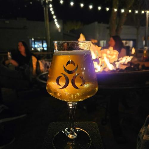 A glass of beer with a logo sits in front of a fire pit, with people socializing in the background under string lights.