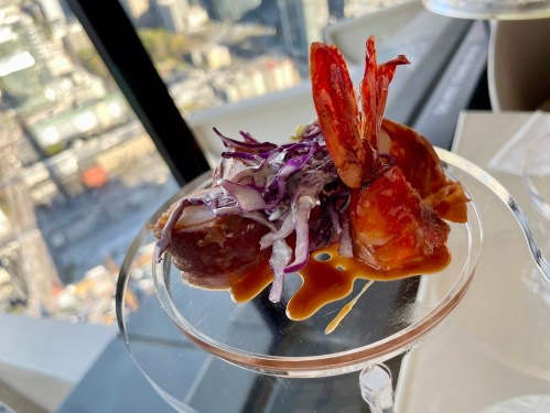 A gourmet dish featuring shrimp, purple cabbage, and a drizzle of sauce, served in a modern glass dish with a city view.