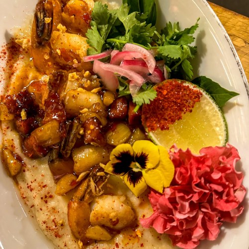 A vibrant dish featuring shrimp, herbs, pickled onions, lime, and edible flowers on a bed of creamy grits.