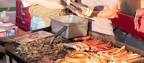 Two people prepare food on a large grill, featuring various sausages and grilled onions.