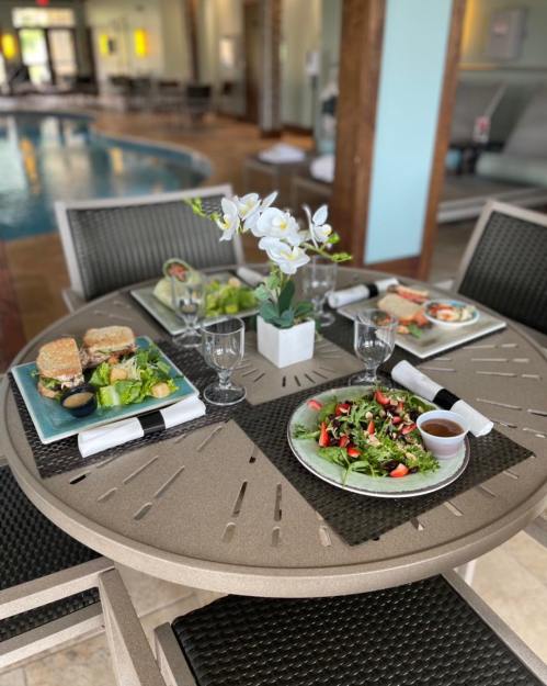 A dining table by a pool with two plates of salad, sandwiches, and drinks, adorned with a flower centerpiece.