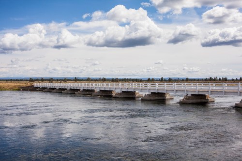 A white wooden bridge spans a calm river under a blue sky with fluffy clouds, surrounded by grassy fields.